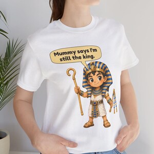 Może przedstawiać: Biały t-shirt z rysunkową ilustracją młodego faraona w niebiesko-złotym pasiatym nakryciu głowy. Postać trzyma złote berło i bicz. Dymek nad postacią zawiera tekst, Mummy says I'm still the king.