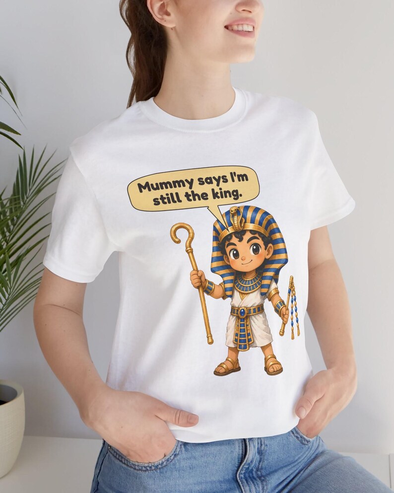 Może przedstawiać: Biały t-shirt z rysunkową ilustracją młodego faraona w niebiesko-złotym pasiatym nakryciu głowy. Postać trzyma złote berło i bicz. Dymek nad postacią zawiera tekst, Mummy says I'm still the king.