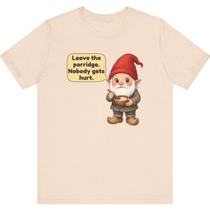 May include: A light beige t-shirt featuring a graphic of a gnome wearing a red hat and brown clothing, holding a wooden bowl with a spoon. A speech bubble next to the gnome contains the text: Leave the porridge. Nobody gets hurt.