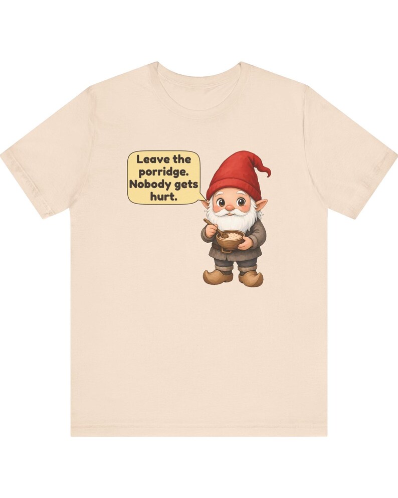 May include: A light beige t-shirt featuring a graphic of a gnome wearing a red hat and brown clothing, holding a wooden bowl with a spoon. A speech bubble next to the gnome contains the text: Leave the porridge. Nobody gets hurt.