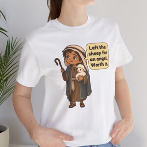 May include: A white t-shirt featuring a cartoon illustration of a shepherd holding a small white lamb and a wooden staff. A yellow speech bubble next to the character contains the text: Left the sheep for an angel. Worth it.