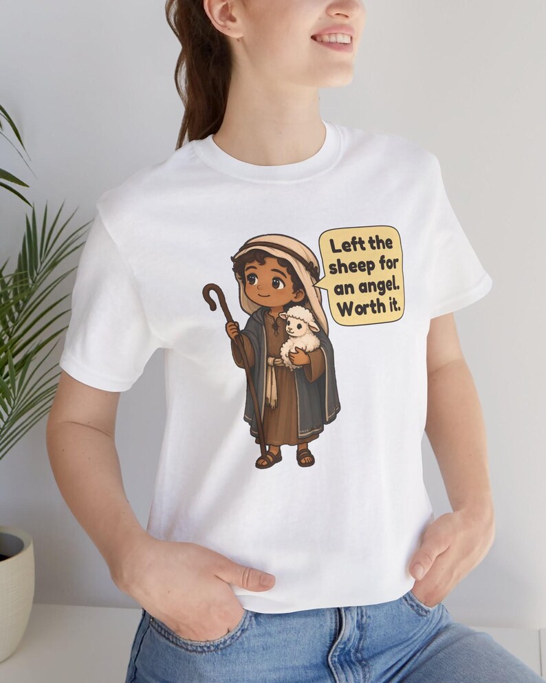 May include: A white t-shirt featuring a cartoon illustration of a shepherd holding a small white lamb and a wooden staff. A yellow speech bubble next to the character contains the text: Left the sheep for an angel. Worth it.