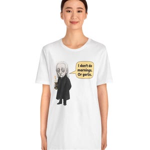 May include: A white short-sleeve t-shirt featuring a graphic of a pale character in a black coat holding a lit candle. A speech bubble next to the character contains the text: I don't do mornings. Or garlic. The design is centered on the chest.