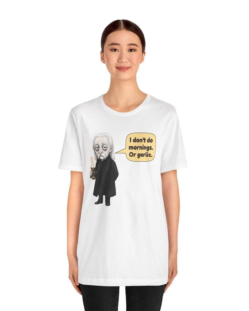 May include: A white short-sleeve t-shirt featuring a graphic of a pale character in a black coat holding a lit candle. A speech bubble next to the character contains the text: I don't do mornings. Or garlic. The design is centered on the chest.
