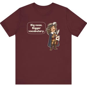 May include: A maroon short-sleeve t-shirt featuring a cartoon illustration of a historical figure with a large nose, wearing a hat and cape, holding a quill and letter. A speech bubble contains the text: Big nose. Bigger vocabulary.
