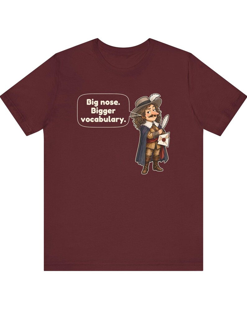 May include: A maroon short-sleeve t-shirt featuring a cartoon illustration of a historical figure with a large nose, wearing a hat and cape, holding a quill and letter. A speech bubble contains the text: Big nose. Bigger vocabulary.