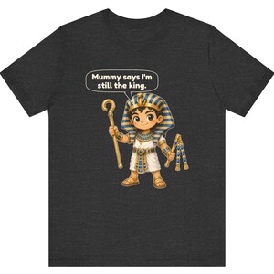 Może przedstawiać: Ciemnoszary t-shirt z rysunkową ilustracją młodego faraona w niebiesko-złotym nakryciu głowy, trzymającego złote berło i bicz. Dymek nad postacią zawiera napis: "Mummy says I'm still the king."