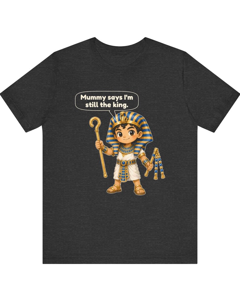 Może przedstawiać: Ciemnoszary t-shirt z rysunkową ilustracją młodego faraona w niebiesko-złotym nakryciu głowy, trzymającego złote berło i bicz. Dymek nad postacią zawiera napis: "Mummy says I'm still the king."