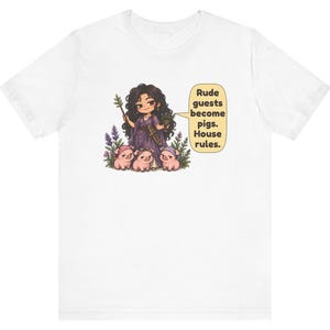 May include: A white t-shirt featuring a graphic of a dark-haired figure in a purple robe holding a wand and a cup, surrounded by three small pink pigs. A speech bubble contains the text: Rude guests become pigs. House rules.