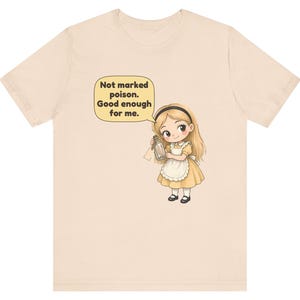 May include: A light beige t-shirt featuring a cartoon illustration of a blonde girl in a yellow dress and white apron holding a small glass bottle. A speech bubble above her contains the text: Not marked poison. Good enough for me.