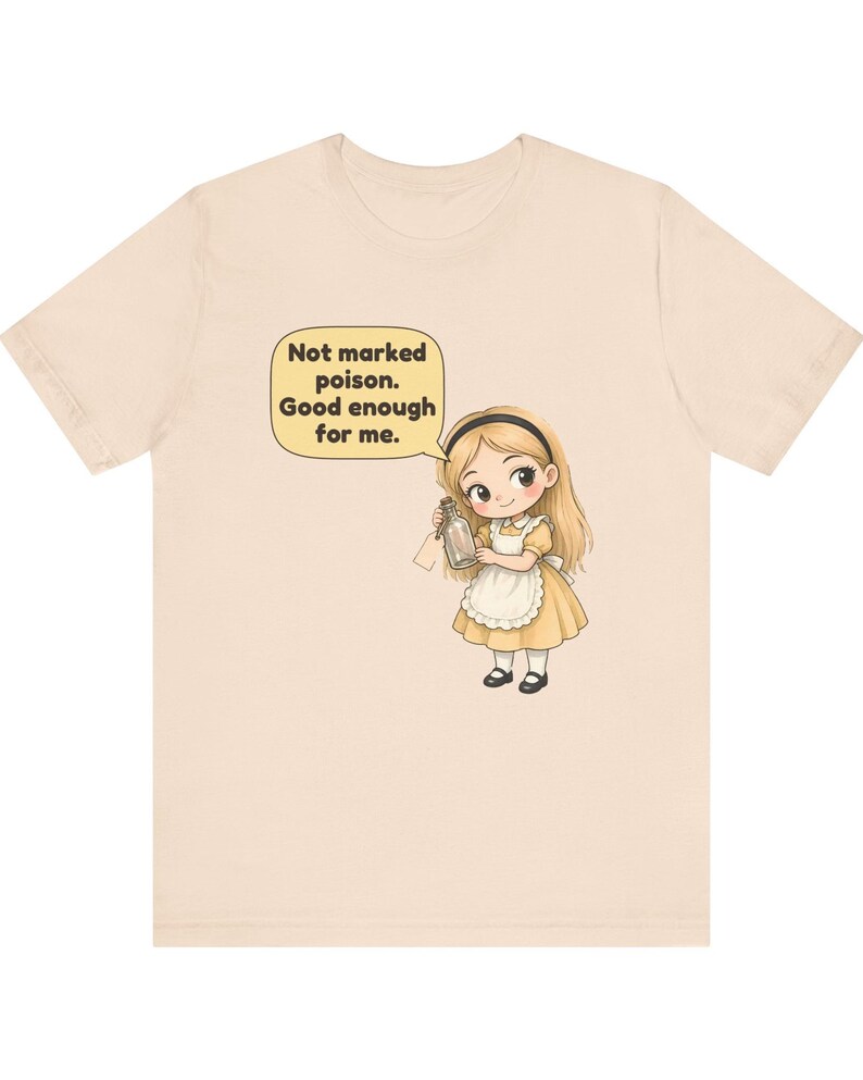 May include: A light beige t-shirt featuring a cartoon illustration of a blonde girl in a yellow dress and white apron holding a small glass bottle. A speech bubble above her contains the text: Not marked poison. Good enough for me.