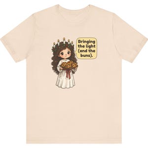May include: A light beige t-shirt featuring a cartoon illustration of a figure wearing a white robe and a candle crown, holding a tray of buns. A speech bubble next to the figure contains the text: Bringing the light (and the buns).