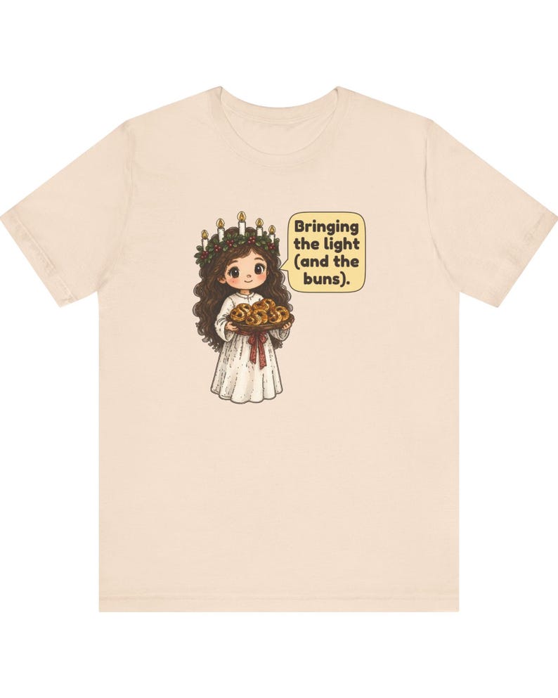 May include: A light beige t-shirt featuring a cartoon illustration of a figure wearing a white robe and a candle crown, holding a tray of buns. A speech bubble next to the figure contains the text: Bringing the light (and the buns).