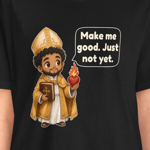 May include: A black t-shirt featuring a cartoon illustration of a figure in gold and white religious vestments and a mitre. The figure holds a brown book and a flaming red heart. A speech bubble contains the text: Make me good. Just not yet.