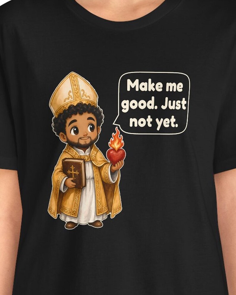 May include: A black t-shirt featuring a cartoon illustration of a figure in gold and white religious vestments and a mitre. The figure holds a brown book and a flaming red heart. A speech bubble contains the text: Make me good. Just not yet.