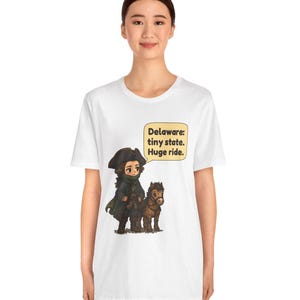 May include: A white short-sleeve t-shirt featuring a cartoon illustration of a historical figure in a dark coat and tricorn hat standing next to a small brown horse. A speech bubble contains the text: Delaware: tiny state. Huge ride.