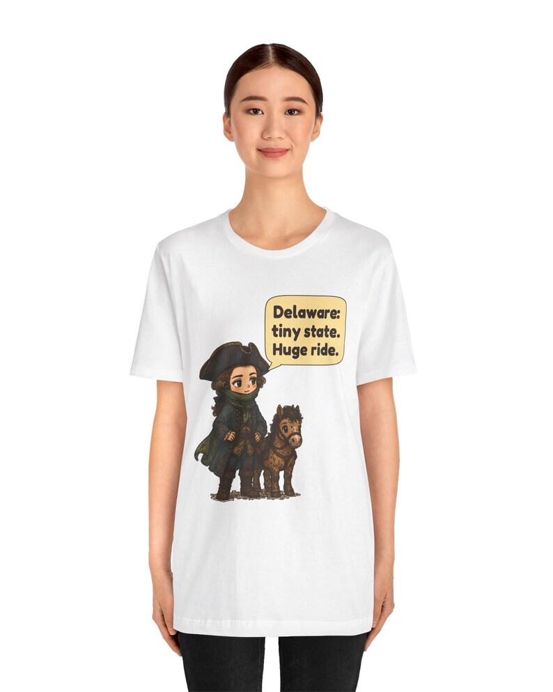 May include: A white short-sleeve t-shirt featuring a cartoon illustration of a historical figure in a dark coat and tricorn hat standing next to a small brown horse. A speech bubble contains the text: Delaware: tiny state. Huge ride.
