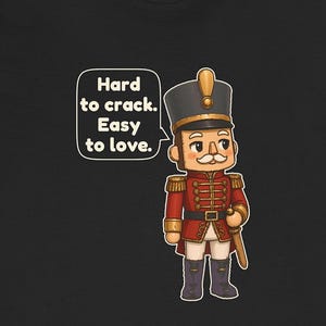 Pu&ograve; includere: Una t-shirt nera con un soldatino schiaccianoci in stile cartoon, con uniforme rossa e dettagli dorati, un cappello alto e baffi bianchi. Un fumetto contiene il testo: Hard to crack. Easy to love.