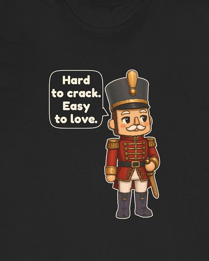 Pu&ograve; includere: Una t-shirt nera con un soldatino schiaccianoci in stile cartoon, con uniforme rossa e dettagli dorati, un cappello alto e baffi bianchi. Un fumetto contiene il testo: Hard to crack. Easy to love.