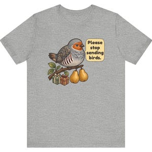 K&ouml;nnte beinhalten: Ein graumeliertes T-Shirt mit einer Grafik eines Vogels, der auf einem Ast mit zwei Birnen und zwei kleinen Geschenken sitzt. Eine Sprechblase neben dem Vogel enth&auml;lt den Text, Please stop sending birds. Der Vogel hat graue und braune Federn.