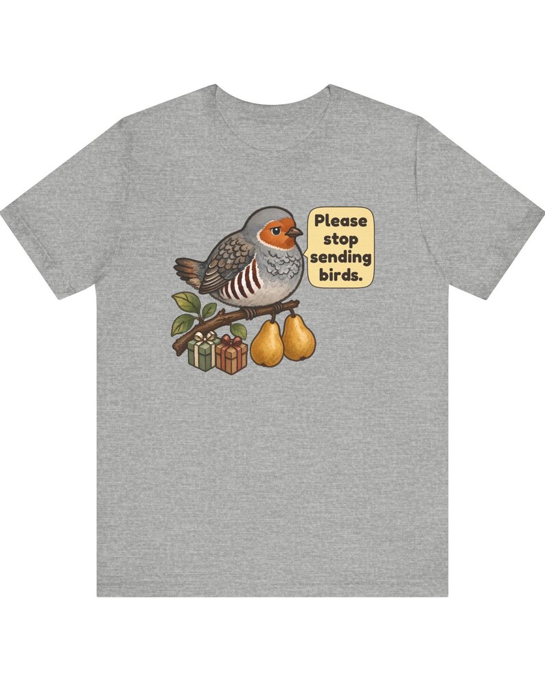 K&ouml;nnte beinhalten: Ein graumeliertes T-Shirt mit einer Grafik eines Vogels, der auf einem Ast mit zwei Birnen und zwei kleinen Geschenken sitzt. Eine Sprechblase neben dem Vogel enth&auml;lt den Text, Please stop sending birds. Der Vogel hat graue und braune Federn.