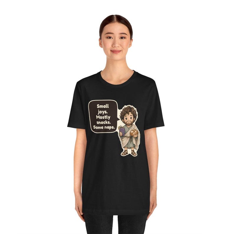 May include: A black short-sleeve t-shirt featuring a cartoon illustration of a bearded figure in a tunic holding grapes and bread. A speech bubble next to the figure contains the text: Small joys. Mostly snacks. Some naps.