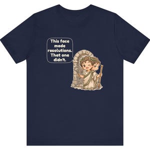 K&ouml;nnte beinhalten: Ein marineblaues T-Shirt mit einer Grafik einer r&ouml;mischen Figur mit zwei Gesichtern, die einen Lorbeerkranz tr&auml;gt und einen Schl&uuml;ssel sowie einen Stab vor einem Torbogen h&auml;lt. Eine Sprechblase enth&auml;lt den Text: This face made resolutions. That one didn't.