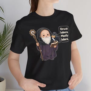 May include: A black t-shirt featuring a cartoon illustration of a bearded monk in a dark robe holding a wooden staff and an open book. A black raven sits on the book, and a speech bubble contains the text: Ora et labora. Mostly labora.