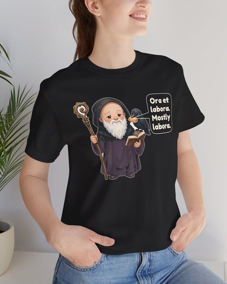 May include: A black t-shirt featuring a cartoon illustration of a bearded monk in a dark robe holding a wooden staff and an open book. A black raven sits on the book, and a speech bubble contains the text: Ora et labora. Mostly labora.