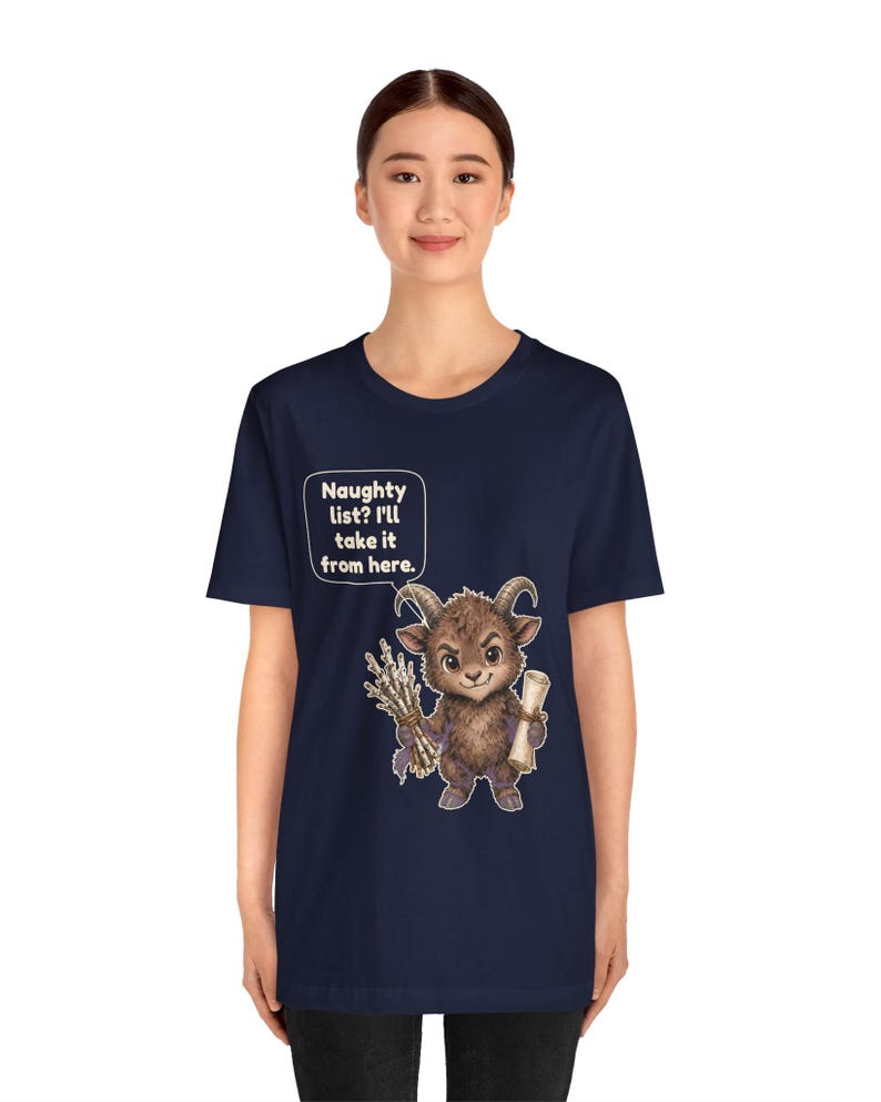 Puede incluir: Una camiseta azul marino de manga corta con el gr&aacute;fico de una criatura marr&oacute;n, peluda y con cuernos que sostiene un manojo de palos y un pergamino. Un bocadillo sobre la criatura contiene el texto: Naughty list? I'll take it from here.