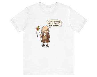 May include: A white t-shirt featuring a cartoon illustration of Benjamin Franklin holding a kite and a key. A speech bubble above the figure contains the text: Kite. Lightning storm. What's your excuse? The design is centered on the chest.