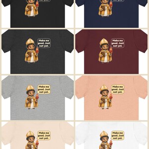 May include: A collection of eight t-shirts in various colors including black, navy, dark gray, maroon, light gray, peach, beige, and white. Each shirt features a cartoon illustration of a saint holding a book and a flaming heart with the text "Make me good. Just not yet."