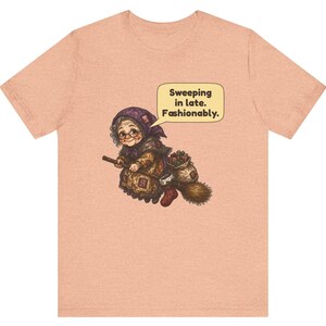 Puede incluir: Una camiseta de manga corta color melocot&oacute;n con un gr&aacute;fico de una persona mayor con pa&ntilde;uelo y gafas montada en una escoba. Un bocadillo sobre la figura contiene el texto: Sweeping in late. Fashionably.