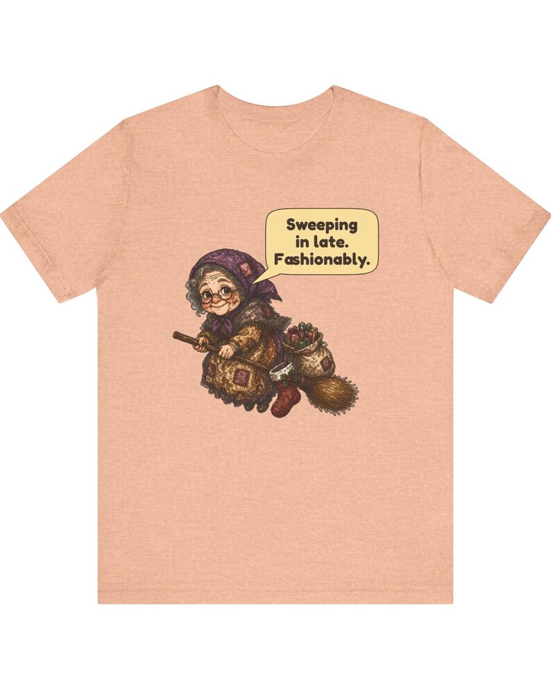 Puede incluir: Una camiseta de manga corta color melocot&oacute;n con un gr&aacute;fico de una persona mayor con pa&ntilde;uelo y gafas montada en una escoba. Un bocadillo sobre la figura contiene el texto: Sweeping in late. Fashionably.