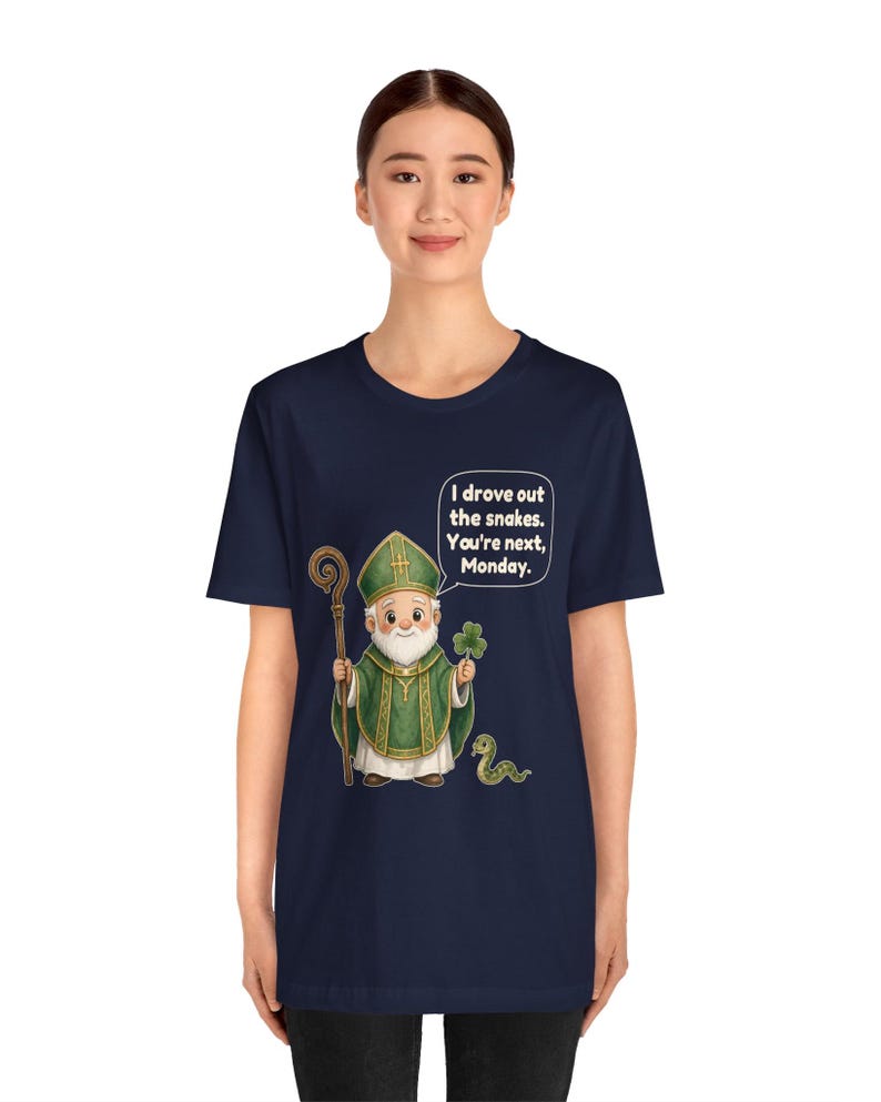 Puede incluir: Una camiseta azul marino de manga corta con una ilustraci&oacute;n de dibujos animados de una figura con vestiduras religiosas verdes que sostiene un bast&oacute;n y un tr&eacute;bol. Una peque&ntilde;a serpiente est&aacute; cerca, y un globo de texto contiene: I drove out the snakes. You're next, Monday.