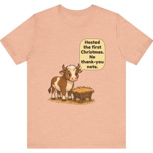 以下が含まれることがあります： ピーチカラーのTシャツ。干し草が入った木製の飼い葉桶の横に立つ茶色と白の子牛が描かれています。子牛の上の吹き出しには「Hosted the first Christmas. No thank-you note.」というテキストが書かれています。