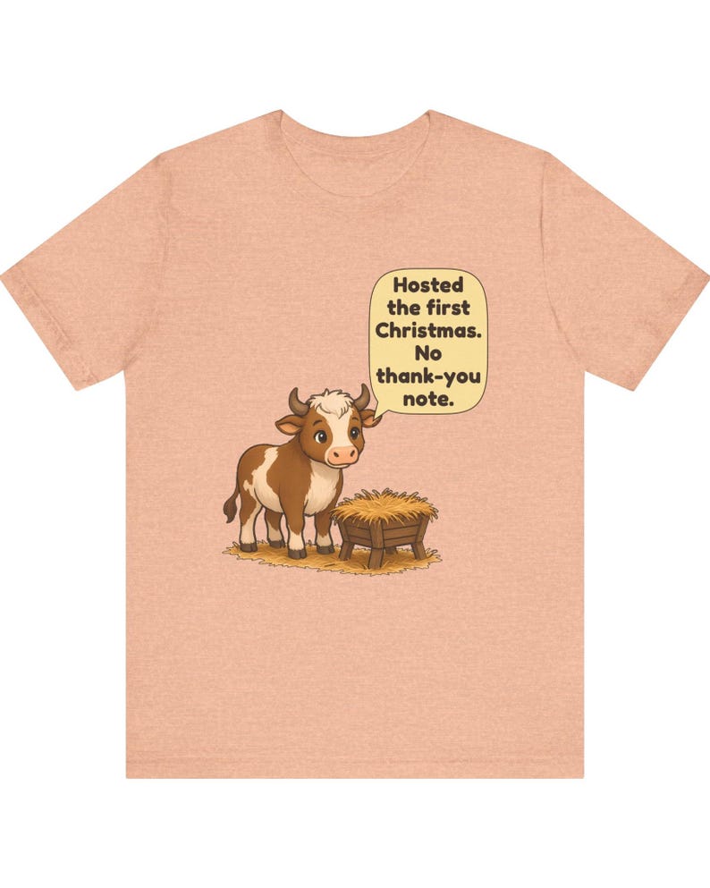 以下が含まれることがあります： ピーチカラーのTシャツ。干し草が入った木製の飼い葉桶の横に立つ茶色と白の子牛が描かれています。子牛の上の吹き出しには「Hosted the first Christmas. No thank-you note.」というテキストが書かれています。