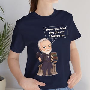 May include: A navy blue t-shirt featuring a cartoon illustration of a man with a white beard and suit holding a stack of books. A speech bubble above him contains the text, "Have you tried the library? I built a few."