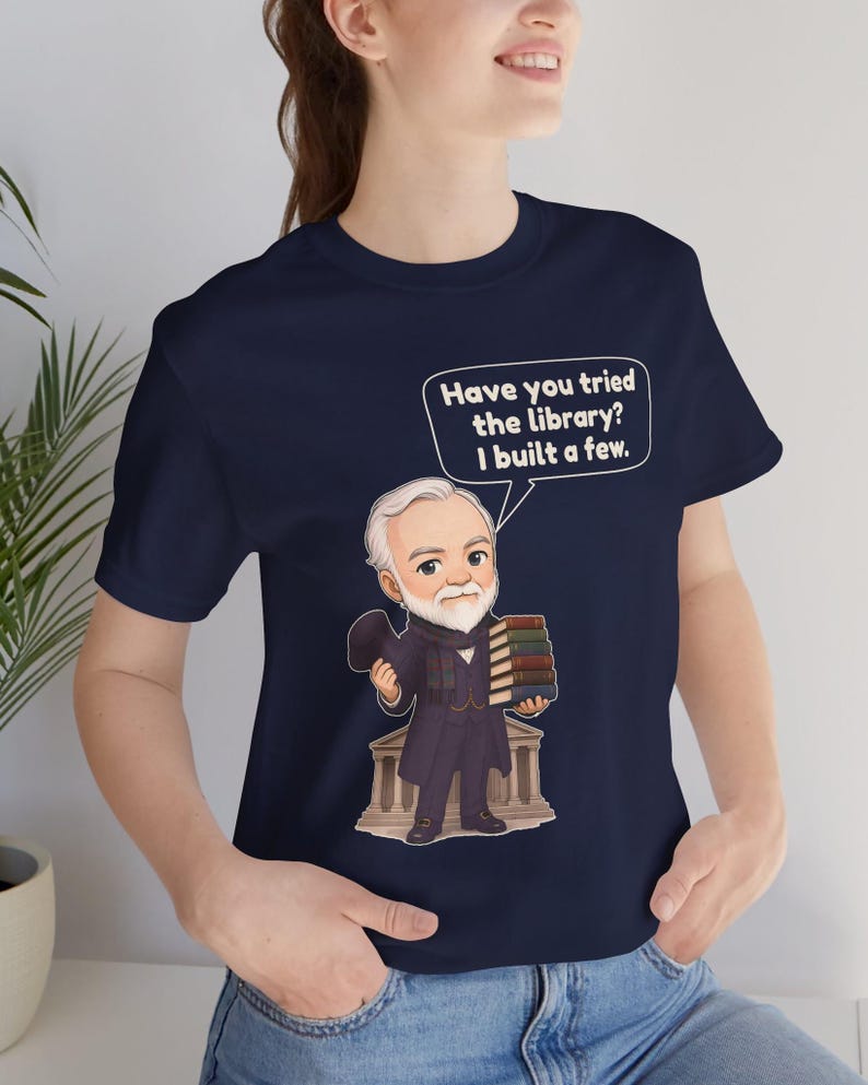 May include: A navy blue t-shirt featuring a cartoon illustration of a man with a white beard and suit holding a stack of books. A speech bubble above him contains the text, "Have you tried the library? I built a few."