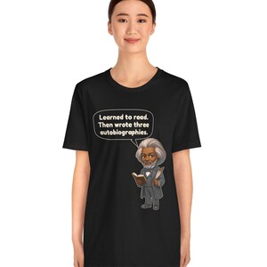 Puede incluir: Una camiseta negra con una ilustraci&oacute;n de dibujos animados de Frederick Douglass sosteniendo un libro y una pluma. Un globo de texto sobre la figura contiene el texto: "Learned to read. Then wrote three autobiographies."