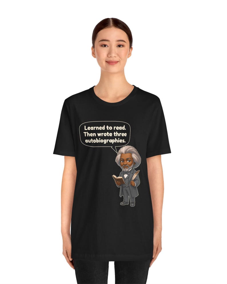 Puede incluir: Una camiseta negra con una ilustraci&oacute;n de dibujos animados de Frederick Douglass sosteniendo un libro y una pluma. Un globo de texto sobre la figura contiene el texto: "Learned to read. Then wrote three autobiographies."