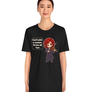 May include: A black t-shirt featuring a cartoon illustration of a red-haired violinist in a dark robe playing a violin. A speech bubble next to the character contains the text: Can't pick a season. Wrote all four.
