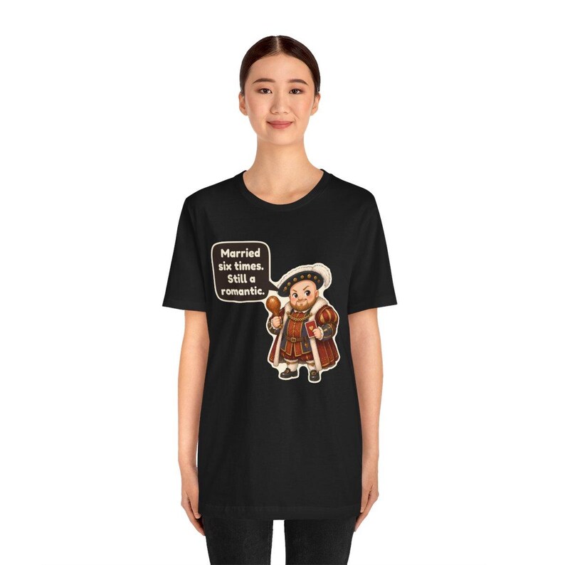 K&ouml;nnte beinhalten: Ein schwarzes T-Shirt mit einer Cartoon-Illustration einer historischen Figur in k&ouml;niglicher Kleidung, die eine H&auml;hnchenkeule und ein kleines rotes Herz h&auml;lt. Eine Sprechblase enth&auml;lt den Text: Married six times. Still a romantic.