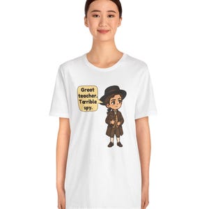 May include: A white short-sleeve t-shirt featuring a cartoon illustration of a person in a brown coat and black hat. A speech bubble next to the character contains the text: Great teacher. Terrible spy. The design is centered on the chest.