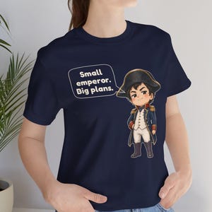 May include: A navy blue t-shirt featuring a cartoon illustration of a historical figure in a military uniform and bicorne hat. Next to the character is a speech bubble containing the white text: Small emperor. Big plans.
