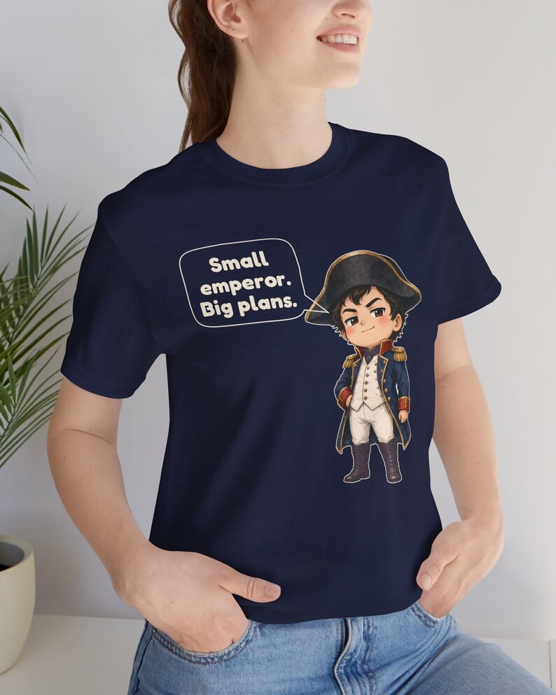 May include: A navy blue t-shirt featuring a cartoon illustration of a historical figure in a military uniform and bicorne hat. Next to the character is a speech bubble containing the white text: Small emperor. Big plans.