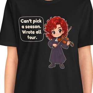 May include: A black t-shirt featuring a cartoon illustration of a red-haired violinist in a dark robe holding a violin. A speech bubble contains the text: Can't pick a season. Wrote all four. The design is centered on the front of the garment.