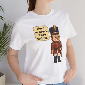 Pu&ograve; includere: Una t-shirt bianca a maniche corte con la grafica di un soldatino schiaccianoci in uniforme rossa e cappello nero. Accanto al soldato c'&egrave; un fumetto giallo contenente il testo nero: Hard to crack. Easy to love.