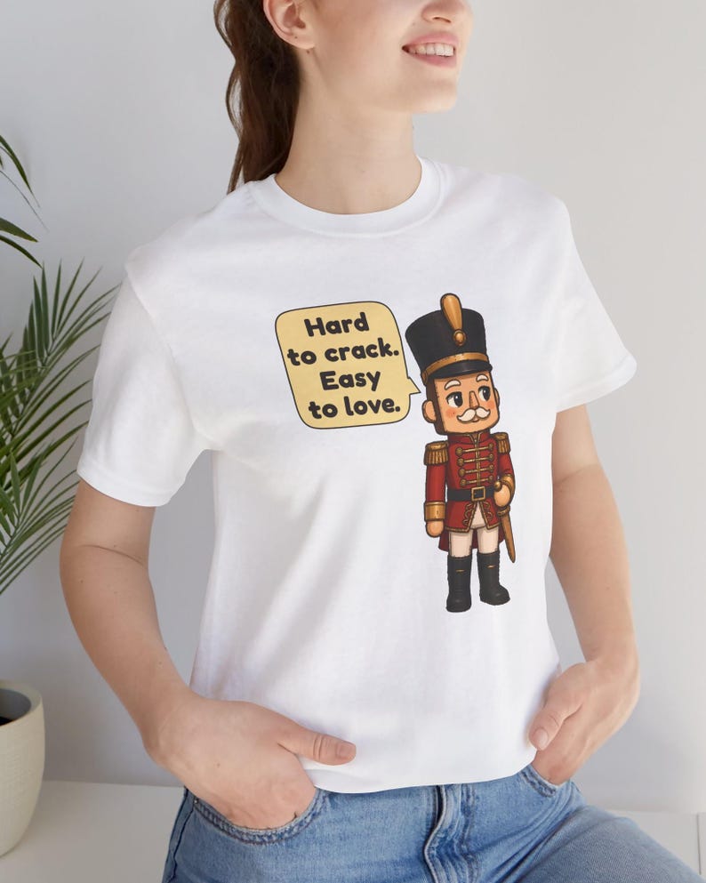 Pu&ograve; includere: Una t-shirt bianca a maniche corte con la grafica di un soldatino schiaccianoci in uniforme rossa e cappello nero. Accanto al soldato c'&egrave; un fumetto giallo contenente il testo nero: Hard to crack. Easy to love.