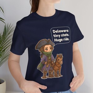 May include: A navy blue t-shirt featuring a graphic of a person in a tricorn hat and historical attire standing next to a small brown horse. A speech bubble above them contains the text: Delaware: tiny state. Huge ride.